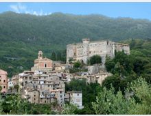 Veduta di un borgo collinare con case in pietra e una chiesa con campanile, dominato da un grande castello fortificato su uno sperone roccioso. Sullo sfondo si estendono pendii montuosi ricoperti di boschi.