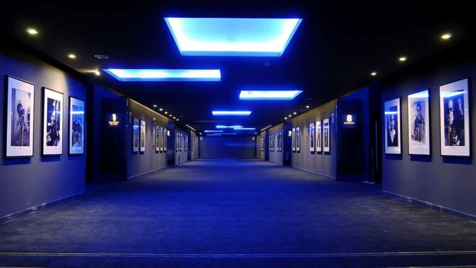 Corridoio interno moderno e ampio, con pareti scure rivestite da fotografie incorniciate ai lati e pavimento in moquette. Il soffitto presenta grandi pannelli luminosi rettangolari con illuminazione blu, creando una prospettiva centrale verso il fondo del passaggio.