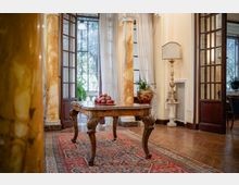 Interno di una villa storica con pavimento in parquet e tappeto persiano, tavolo in legno intagliato al centro e colonne in marmo. Grandi porte e finestre vetrate con tende leggere affacciano sul giardino; a lato si vede una lampada da terra e un vaso con pianta.