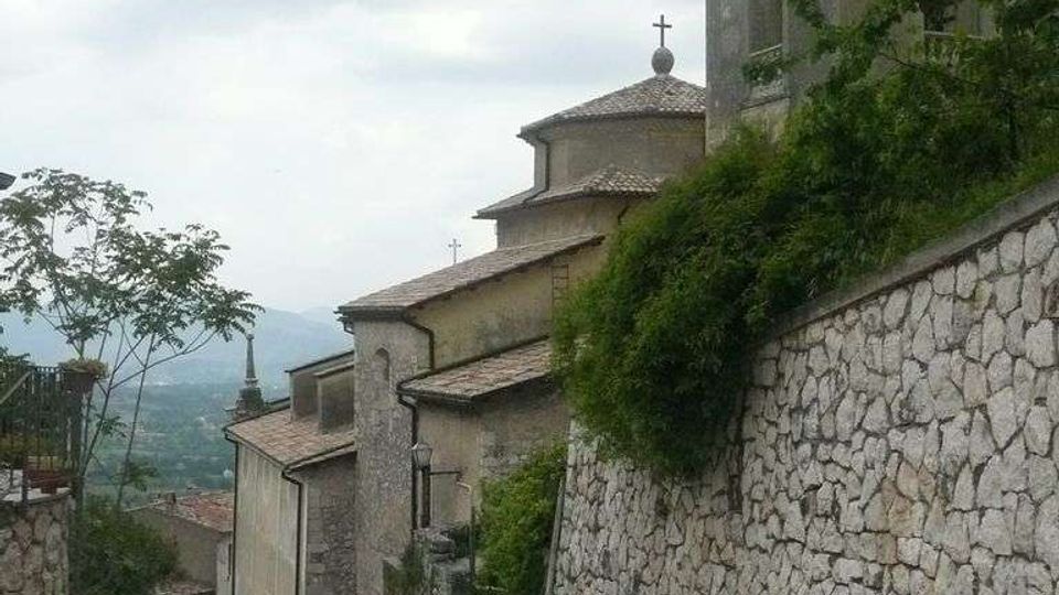 Strada in pendenza lastricata in pietra in un borgo collinare, affiancata da un alto muro in pietra e vegetazione. Sul lato destro si vede una chiesa in muratura con tetti in coppi e un campanile, con montagne sullo sfondo.