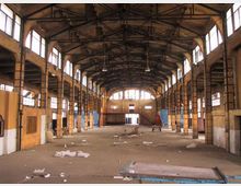 Interno di un grande capannone industriale dismesso, con struttura metallica a capriate sul soffitto e alte finestre laterali che illuminano lo spazio. Pavimento in cemento quasi vuoto con detriti sparsi e pareti in muratura con pilastri e impalcati laterali.