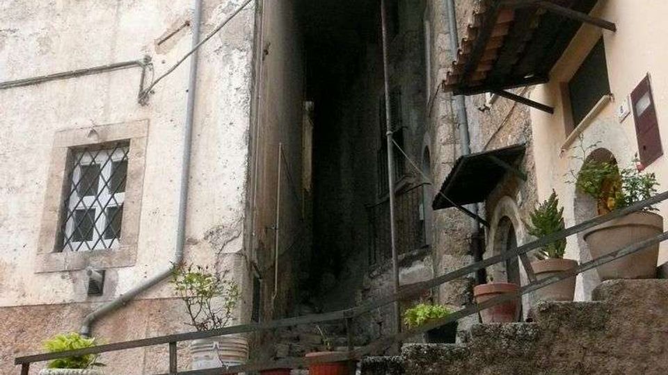 Vicolo stretto tra edifici in muratura e intonaco invecchiato, con una scalinata ripida che sale in un passaggio coperto tra le case. In primo piano un muretto in pietra con ringhiera e vasi di piante, e finestre con persiane e grate sulle facciate.