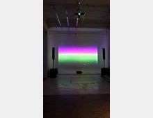 Interno di uno spazio espositivo industriale con pavimento in cemento e soffitto a travi, con un proiettore sospeso. Su una parete bianca è proiettata un’immagine rettangolare a bande colorate viola e verde, affiancata da due altoparlanti su supporti.