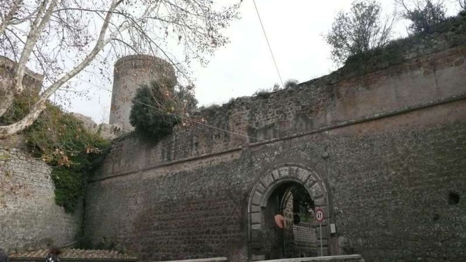 Mura di fortificazione in mattoni con grande arco d’ingresso in pietra e passaggio sottostante, affiancate da un torrione cilindrico sullo sfondo. Davanti si vede una strada con muretto curvo e alberi spogli lungo il lato sinistro.