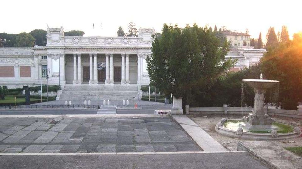 Veduta di un grande edificio monumentale in stile neoclassico con ampia scalinata centrale e portico colonnato. In primo piano c’è una piazza lastricata e, sulla destra, una fontana circolare, con alberi e giardini ai lati.