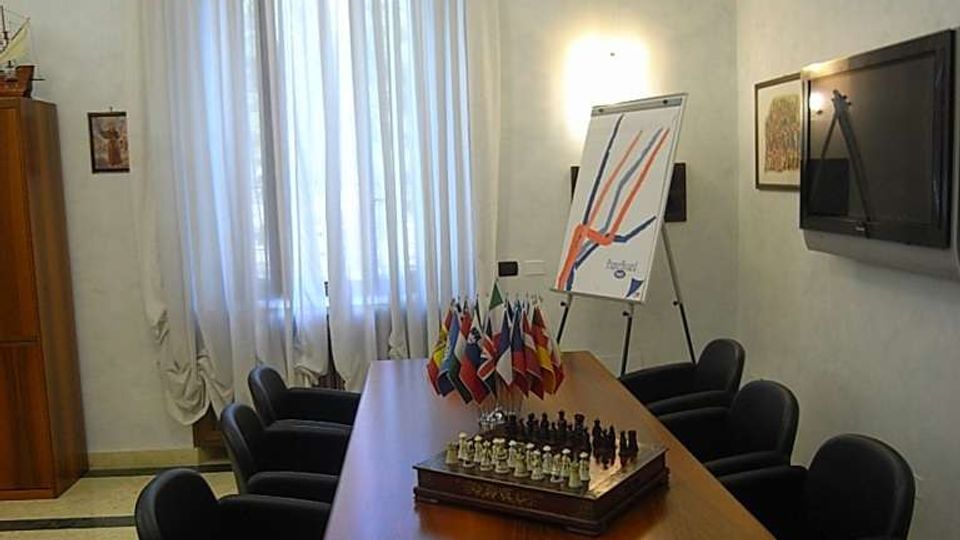 Sala riunioni con lungo tavolo in legno circondato da poltroncine scure, con al centro una scacchiera e piccole bandiere. Sul fondo una grande finestra con tende leggere, mentre sulle pareti ci sono un televisore e un cavalletto con lavagna a fogli mobili.