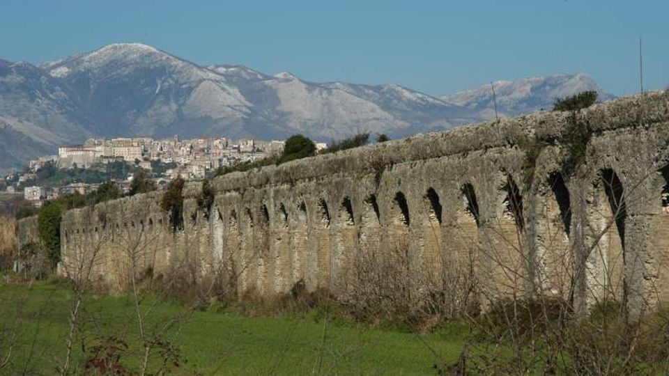 Lungo acquedotto in pietra con una serie di archi ripetuti, che attraversa un prato con vegetazione bassa. Sullo sfondo si vedono un centro abitato su una collina e alte montagne sotto un cielo sereno.