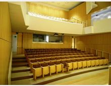 Interno di un auditorium moderno con pareti rivestite in legno chiaro e file di sedute in legno disposte su gradinate. In alto si vede un balcone con parapetto e una piccola cabina tecnica affacciata sulla sala, illuminata da faretti a soffitto.