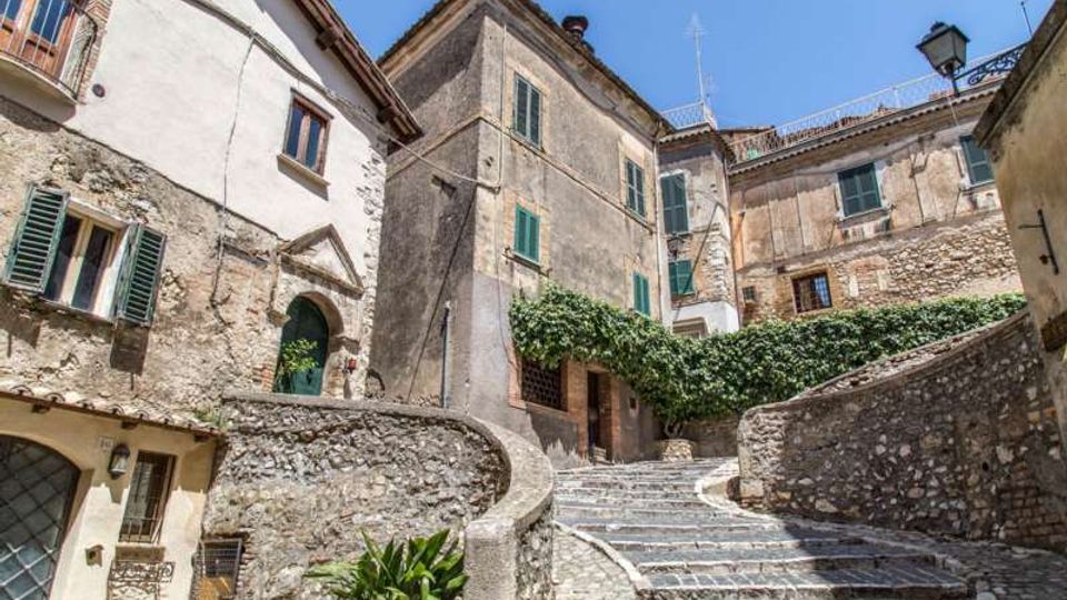 Scalinata in pietra che sale tra edifici storici con facciate intonacate e muri in pietra, con finestre e persiane verdi. Muretti curvi delimitano i gradini e una siepe di vegetazione corre lungo il lato destro sotto un cielo sereno.