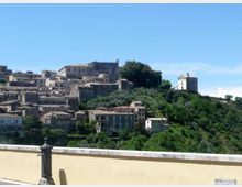 Veduta panoramica di un borgo collinare con case in pietra e tetti in coppi disposte a terrazze tra uliveti e vegetazione. Sulla cresta si distinguono un edificio storico e una piccola chiesa isolata sul pendio, sotto un cielo sereno.