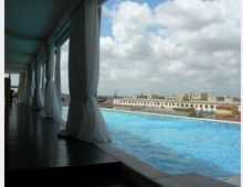 Terrazza con piscina a sfioro affiancata da una passerella in legno coperta, con tende bianche sospese lungo il lato. Sullo sfondo si vede lo skyline di edifici bassi della città sotto un cielo parzialmente nuvoloso.