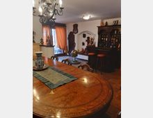Sala da pranzo in stile tradizionale con grande tavolo ovale in legno intarsiato, lampadario e pavimento in cotto. Sullo sfondo si vedono un angolo bar con sgabelli, una vetrina con bottiglie e oggetti decorativi, e una porta-finestra con tende arancioni che conduce a un balcone.