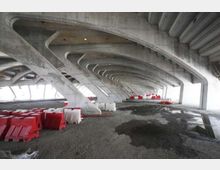 Spazio sotto un grande viadotto in cemento armato, con una serie di archi e pilastri inclinati ripetuti in prospettiva. Sul pavimento in ghiaia e asfalto con pozzanghere sono presenti barriere stradali in plastica rosse e bianche disposte lungo i lati.