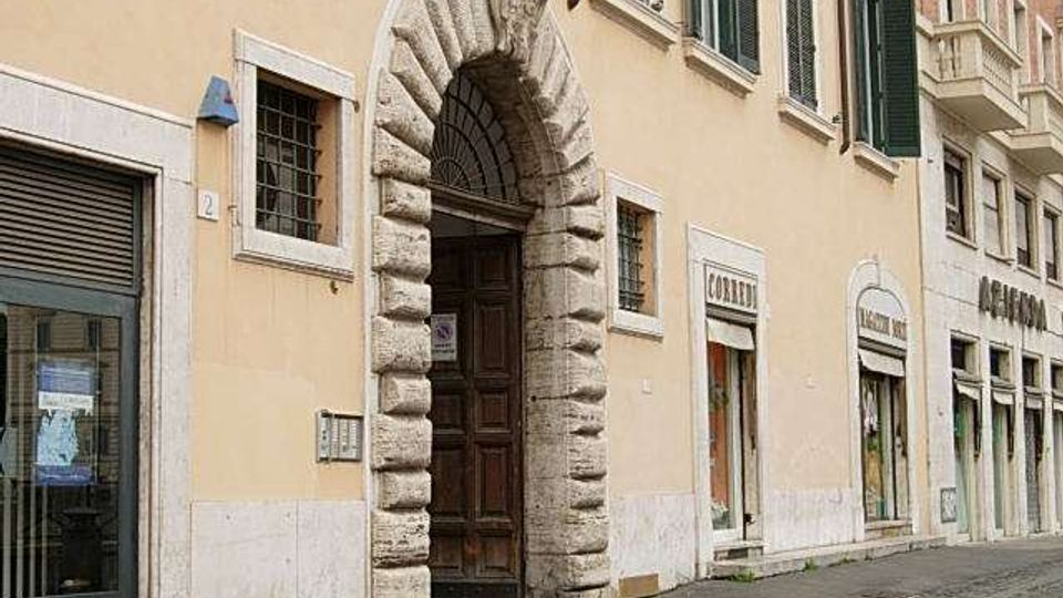 Facciata di edificio storico in una via urbana, con grande portale ad arco in pietra bugnata e porta in legno scuro. Al piano strada si vedono vetrine di negozi e finestre con inferriate, mentre ai piani superiori ci sono finestre con persiane verdi.