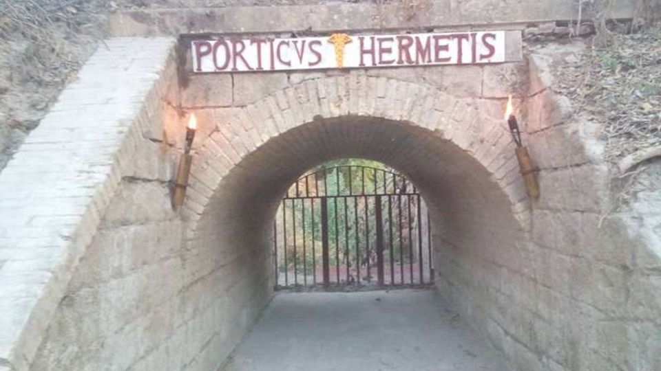 Ingresso a tunnel ad arco in pietra con volta in mattoni e pavimento in cemento, sormontato dall’insegna “PORTICVS HERMETIS”. Ai lati dell’arco sono fissate due torce e in fondo si vede un cancello in ferro che chiude il passaggio verso un’area alberata.