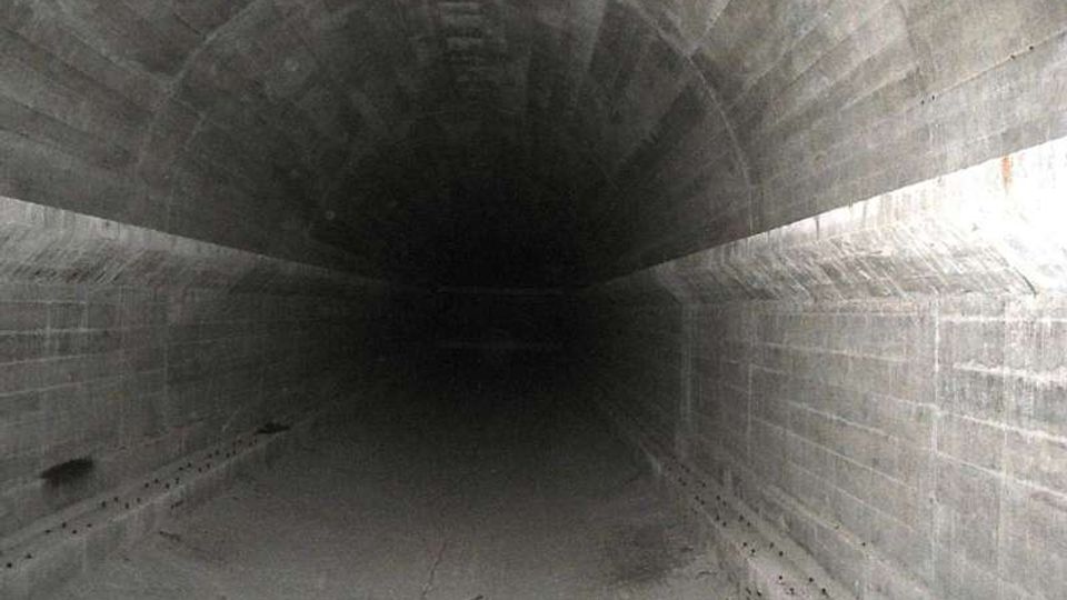 Interno di un tunnel sotterraneo in cemento con volta ad arco e pareti scanalate, che si estende in profondità verso un’area buia. Il pavimento è in cemento e presenta crepe e segni di usura lungo i bordi.