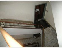 Vano scala interno di un edificio, visto dall’alto, con gradini in cemento, corrimano in legno e ringhiera metallica. Sul pianerottolo si vedono pareti chiare, pavimento in pietra/mosaico e una porta scura con cornice in marmo.