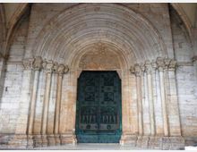 Portale monumentale in pietra di una chiesa, con grande arco a tutto sesto a stadi concentrici e colonne laterali con capitelli scolpiti. Al centro si trova una pesante porta doppia in metallo scuro decorata con motivi geometrici in rilievo.