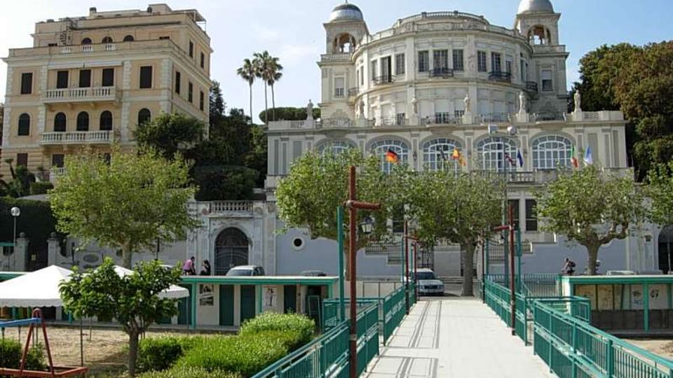 Viale pedonale con ringhiere verdi che conduce a un grande edificio storico in stile neoclassico, con facciata curva, logge ad archi e due torrette con cupole. Intorno si vedono alberi, giardini e un altro palazzo chiaro sulla sinistra, sullo sfondo di una collina.