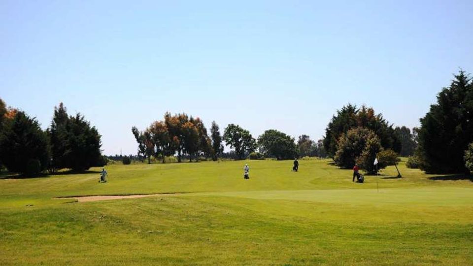 Ampio campo da golf con prato rasato e ondulato, delimitato da alberi e cespugli lungo i bordi. In primo piano si vede un bunker di sabbia e, sulla destra, una bandierina che segna il green sotto un cielo sereno.