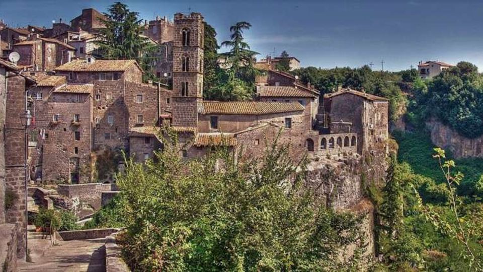 Borgo storico con edifici in pietra dai tetti in coppi, affacciato su una scarpata rocciosa sopra una valle boscosa. Al centro si distingue una torre campanaria e, lungo il bordo del dirupo, un edificio con loggiato ad archi.