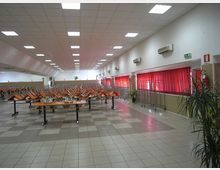 Ampia mensa o sala refettorio con pavimento a piastrelle e controsoffitto con pannelli luminosi, con tavoli lunghi e sedie capovolte sopra. Lungo la parete destra ci sono finestre con tende rosse, climatizzatori a parete e carrelli metallici.