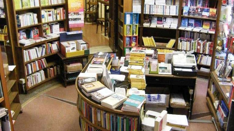 Interno di una libreria con alte scaffalature in legno piene di libri e una postazione centrale curva con pile di volumi e attrezzature da cassa. Sul fondo si vede una porta con l’insegna “Narrativa e poesia” che conduce a un corridoio con scala.