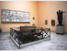 Sala museale con pareti color ocra e pavimento in pietra, con al centro un grande modello circolare recintato da una struttura metallica. Sulle pareti sono esposti un lungo rilievo marmoreo e alcune lapidi, con una grande statua scura su piedistallo in un angolo.
