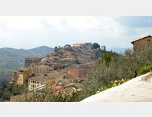 Panorama di un borgo collinare con case in pietra dai tetti in coppi, disposto a gradoni sul pendio e dominato da edifici storici e una torre campanaria. In primo piano si vedono vegetazione e un muretto, con colline e montagne sullo sfondo sotto un cielo nuvoloso.