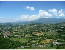 Veduta panoramica di una valle collinare con campi coltivati, boschi e piccoli borghi sparsi. Sullo sfondo si vedono montagne verdi sotto un cielo azzurro con nuvole.
