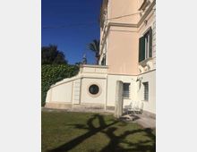 Facciata esterna di un edificio in stile classico color crema e bianco, con cornici decorative, persiane verdi e una finestra circolare in un muretto laterale. In primo piano c’è un prato con ombre di palme; a lato si vedono una panchina metallica e finestre con inferriate.