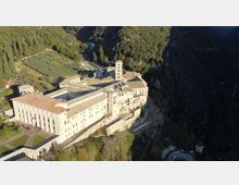 Veduta aerea di un grande complesso monastico in pietra chiara con tetti in coppi rossi, chiostro interno e campanile, costruito sul bordo di una scarpata. L’edificio è circondato da boschi e campi coltivati, con strade e piccoli edifici rurali nelle vicinanze.