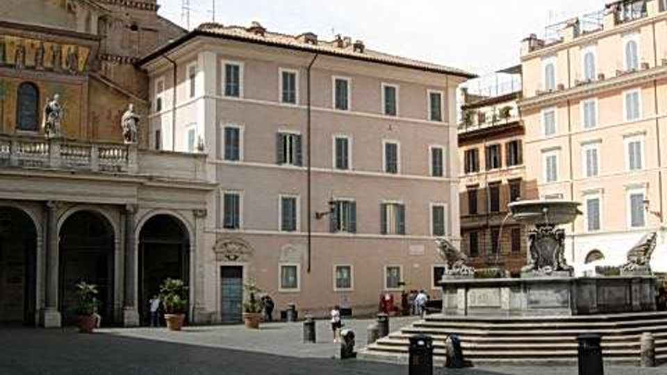 Piazza lastricata in pietra con edifici storici dai toni chiari e una torre campanaria con grande orologio sulla facciata. Al centro si vede una fontana monumentale rialzata con vasca e sculture, circondata da gradini e paletti.