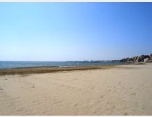 Ampia spiaggia sabbiosa sul mare con linea di battigia e acqua calma sotto un cielo sereno. Sullo sfondo, a destra, si vede la costa con edifici lungo il litorale e un promontorio all’orizzonte.