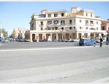 Ampia piazza asfaltata con parcheggio e auto davanti a un grande edificio d’angolo a più piani, con facciata chiara, balconi e portici al piano terra con negozi. Sullo sfondo si vedono altri palazzi lungo la strada e alcune palme ai margini.