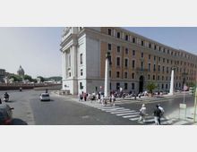 Incrocio urbano con ampio attraversamento pedonale davanti a un grande edificio storico in muratura chiara con facciata lunga e finestre regolari. Strada lastricata con traffico, marciapiedi e segnaletica; sullo sfondo si vede una cupola e un ponte in lontananza.