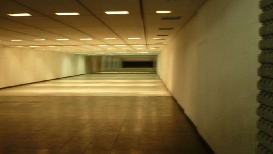 Ampio corridoio sotterraneo con pavimento piastrellato e pareti bianche, illuminato da una griglia di luci rettangolari incassate nel soffitto. La prospettiva conduce verso un’area più buia in fondo, con una breve scalinata e pannelli o vetrate.