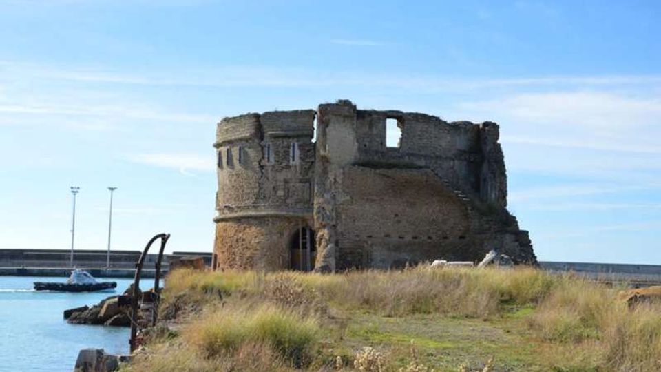 Rovine di una torre o fortificazione in pietra su un promontorio erboso, affacciata su un porto con acqua calma e una diga frangiflutti sullo sfondo. La struttura è cilindrica e parzialmente crollata, con aperture murate e muri irregolari.