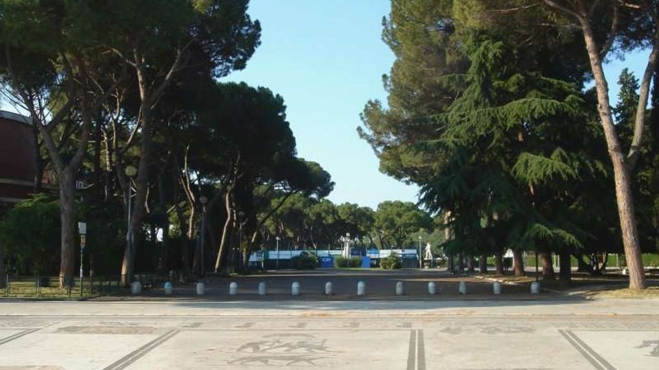 Ampio piazzale pavimentato con motivi decorativi, delimitato da una fila di dissuasori e circondato da alti pini e altre conifere. Sullo sfondo si intravede un’area verde con strutture e cartelli, sotto un cielo sereno.