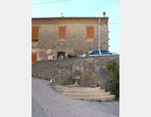 Fontana in pietra con vasca su gradini semicircolari lungo una strada in pendenza, addossata a un muro di contenimento in muratura. Sullo sfondo si vede una casa in pietra a due piani con persiane in legno e tetto in coppi.