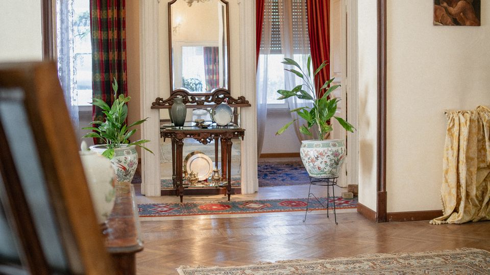 Interno di una casa in stile classico con pavimento in parquet e grande tappeto; un ampio varco incorniciato in legno conduce a un salotto con lampadario, consolle con specchio e due vasi con piante. Ai lati si vedono porte con tende rosse e a fantasia, e piccoli quadri alle pareti.