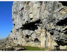 Facciata in pietra di una piccola chiesa incastonata alla base di un’imponente parete rocciosa a strapiombo, con sporgenze naturali che creano ombra. In primo piano si vede un prato e una passerella con staccionata in legno, sullo sfondo un cielo azzurro e il paesaggio montano.