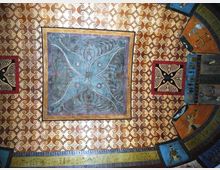 Soffitto decorato con motivi geometrici ripetuti in oro e crema e un grande riquadro centrale blu con disegno simmetrico a quattro petali. Ai lati si vedono pannelli quadrati e fasce curve dipinte con ornamenti e figure stilizzate lungo il bordo.