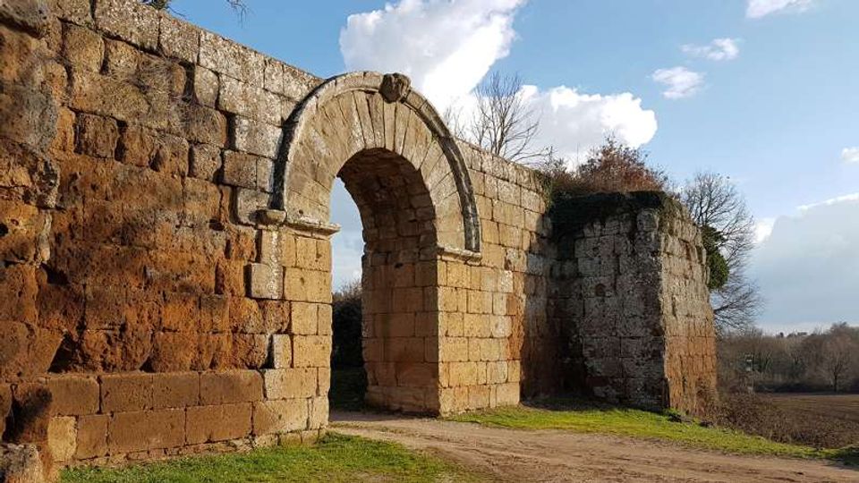 Resti di una struttura in pietra con un grande arco a tutto sesto inserito in un muro antico. Davanti passa un sentiero sterrato su un prato, con campagna e alberi spogli sullo sfondo sotto un cielo con nuvole.