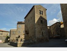 Torre in pietra in un borgo storico, con piccole finestre e apertura a bifora nella parte superiore. Intorno si vedono edifici in muratura e una piazzetta con pavimentazione in pietra sotto un cielo sereno.