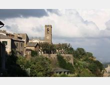 Veduta di un borgo collinare con edifici in pietra e un campanile con orologio che svetta al centro. In primo piano si vede vegetazione fitta e una terrazza murata con alberi potati, sullo sfondo cielo nuvoloso e paesaggio in lontananza.