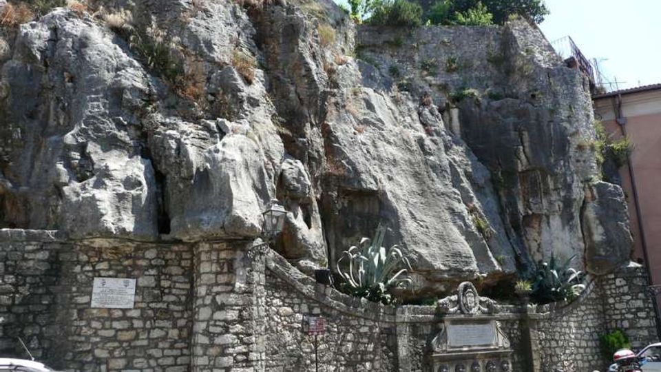 Rupe rocciosa a picco sopra un alto muro in pietra, con vegetazione mediterranea e palme sulla sommità. In basso si vede un piccolo spazio urbano con muretto, lapide/monumento in pietra, lampione, vasi e auto parcheggiate lungo la strada.
