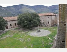 Cortile erboso di un complesso in pietra con edifici dai tetti in tegole, un grande albero centrale e un pozzo circolare su un’area di ghiaia. Sullo sfondo si vedono colline e montagne boscose.