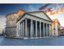 Tempio romano in pietra con grande portico a colonne e frontone triangolare, affiancato da una rotonda muraria sul retro. L’edificio si trova in una piazza lastricata, circondato da palazzi cittadini sotto un cielo al tramonto.