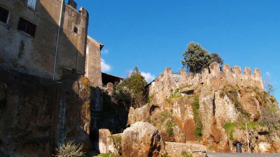 Complesso storico in pietra su uno sperone roccioso, con edifici e una cinta muraria merlata che seguono il profilo della rupe. In primo piano si vede una strada asfaltata in salita con muretto e piccoli spazi verdi, sotto un cielo limpido.